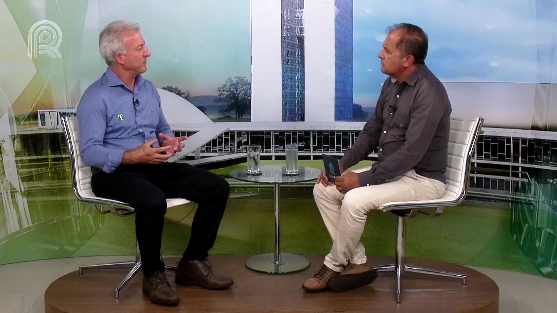 Two men are seated in chairs, engaged in a conversation, likely on a program from Canal Rural in Brazil, given the backdrop. The man on the left gestures with his hands, while the other holds a small object.

