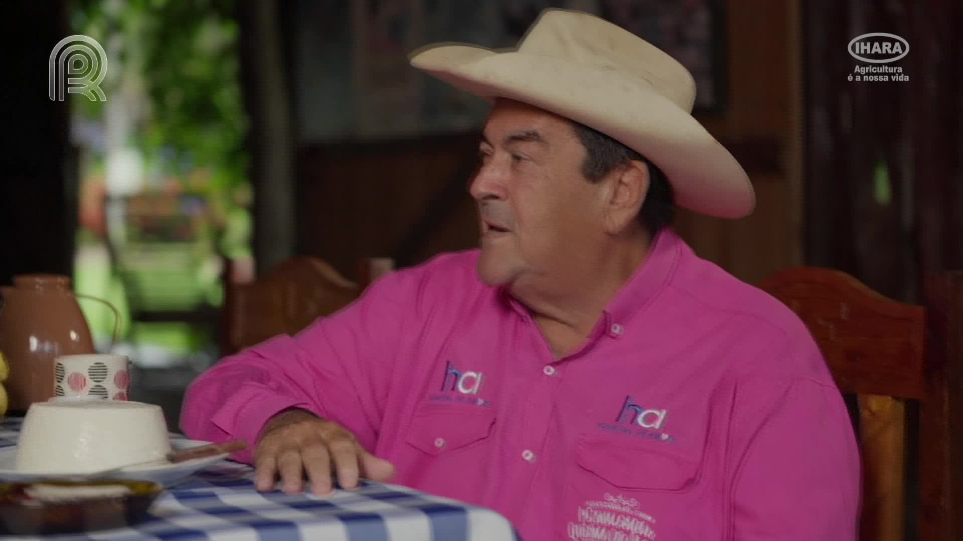 A man in a pink shirt and cowboy hat gestures while seated at a table. A plate of food and a coffee cup sit before him on a blue-and-white checkered tablecloth.
