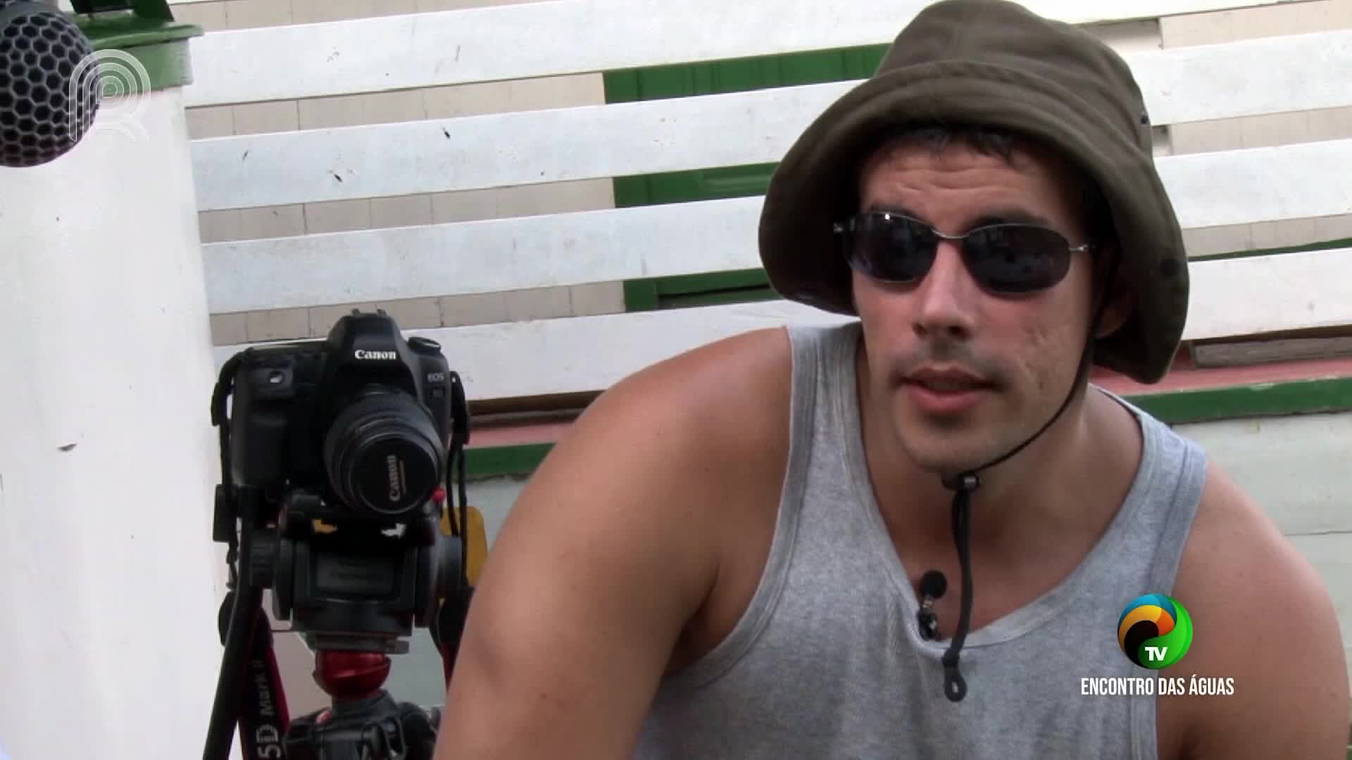 A man in a hat and sunglasses is speaking, a microphone clipped to his shirt. A Canon camera on a tripod sits nearby, ready to capture the scene for Canal Rural in Brazil.
