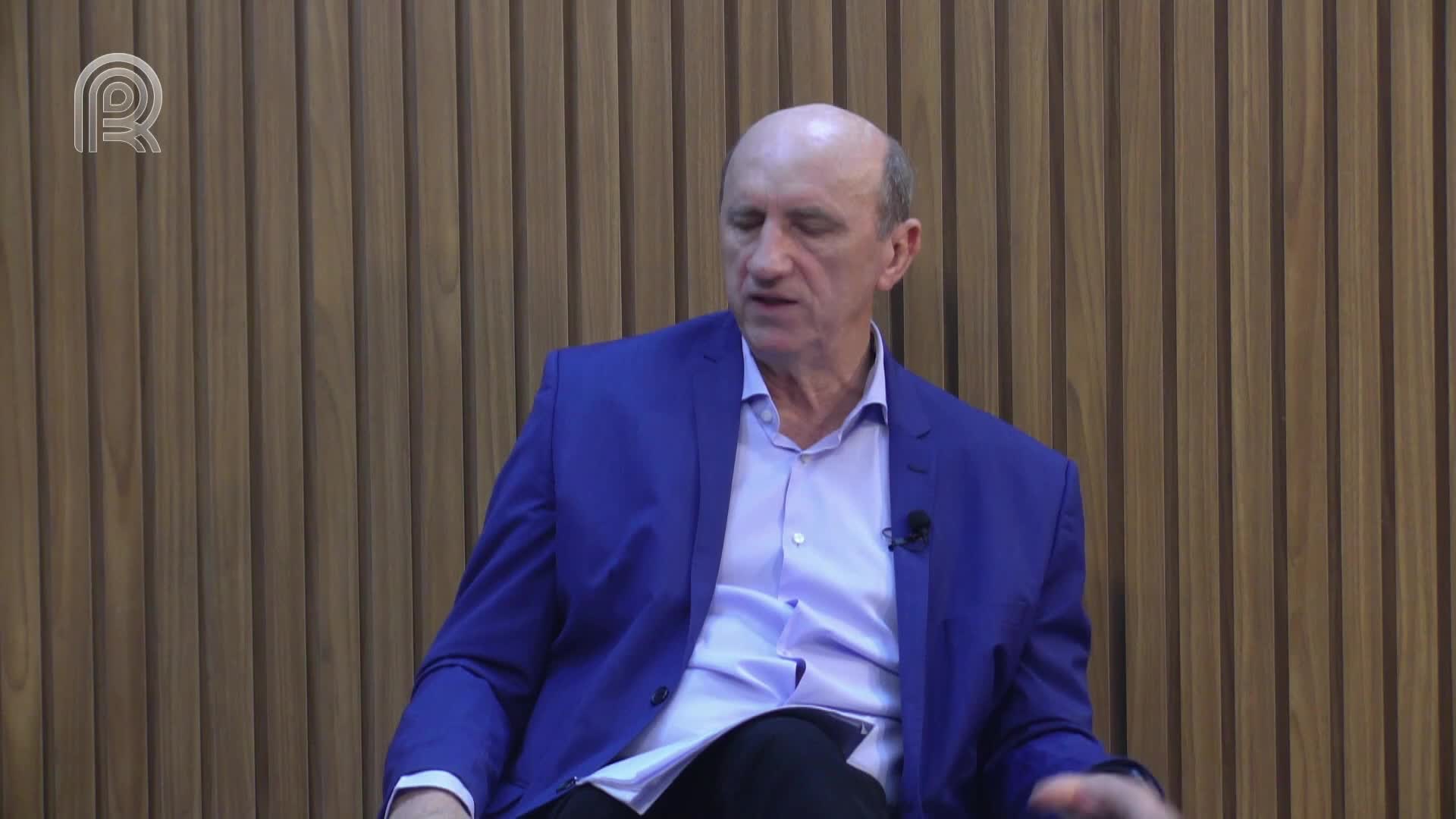 A man in a blue blazer sits, speaking, against a backdrop of vertical wooden panels. A small microphone is clipped to his shirt.

