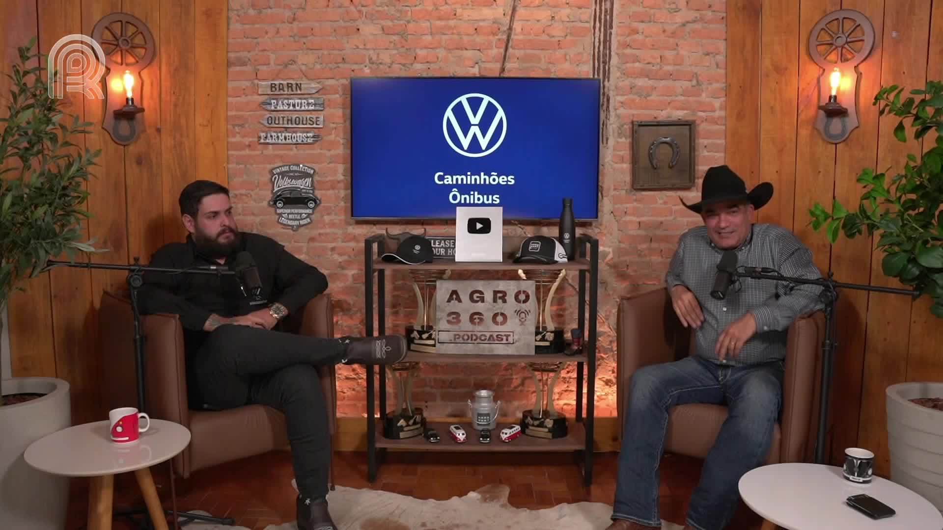 Two men sit in armchairs, microphones positioned in front of them. One is wearing a cowboy hat, and behind them, a screen displays a Volkswagen logo, likely for a broadcast on Canal Rural in Brazil.
