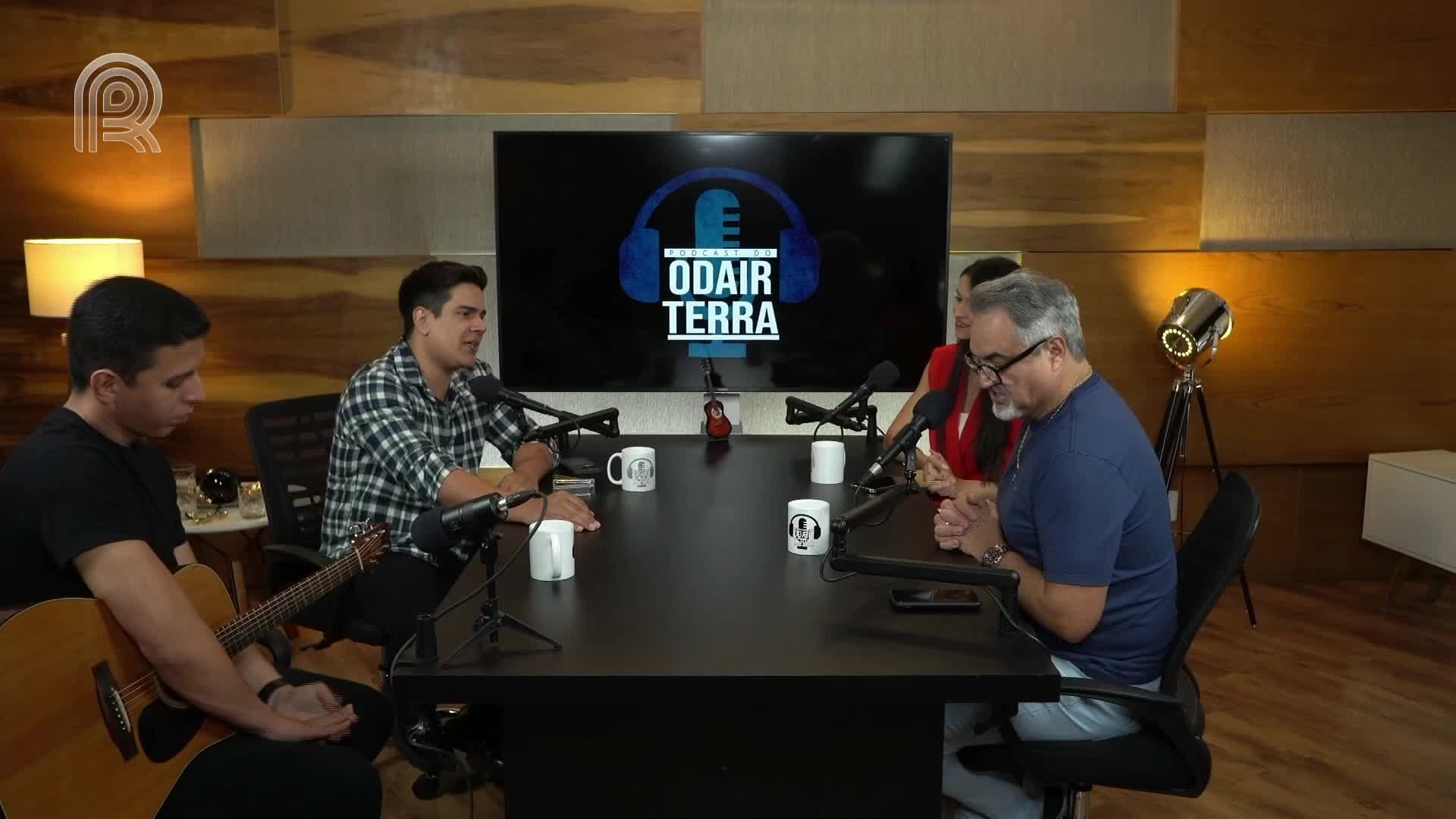 Four people sit around a table, microphones in front of them, the logo of "Odair Terra" on the screen behind. A man strums a guitar, while others speak into their mics, likely for a Canal Rural podcast in Brazil.
