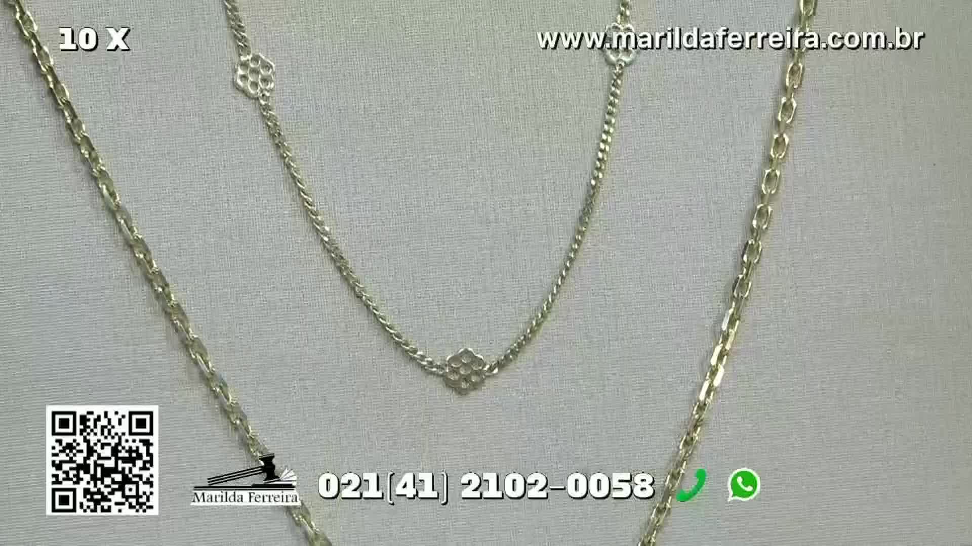 Three silver necklaces hang against a textured white backdrop. The necklaces, with their different chain styles and geometric charms, are likely being presented on a broadcast from Brazil.
