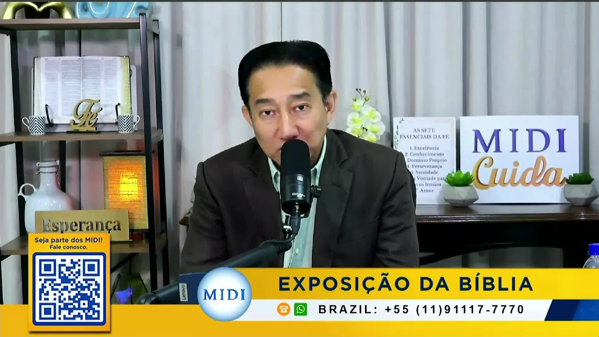 A man in a brown jacket speaks into a microphone, with a backdrop of shelves holding books and decorative items. A banner at the bottom advertises an exposition in Brazil, with a phone number.
