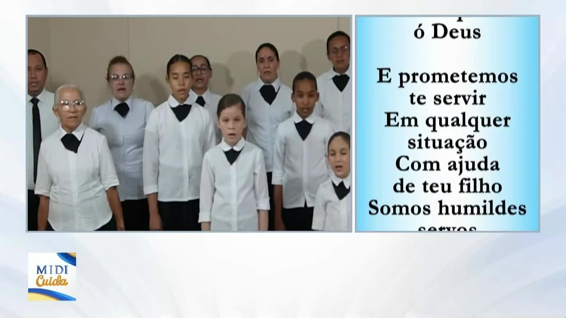 A group of people in white shirts and black ties stand in a row, singing. Beside them, lyrics in Portuguese scroll on a blue background.
