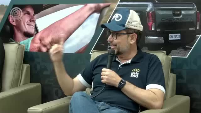 A man in a blue cap and glasses gestures with his left hand while holding a microphone. Behind him, a screen shows a man in a car and a Chevrolet truck.
