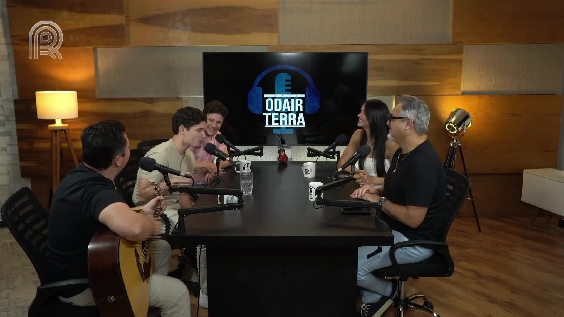 A group of people are seated around a table, microphones in front of them, as if they're on a Canal Rural podcast. One man strums a guitar while others appear to be talking and listening.
