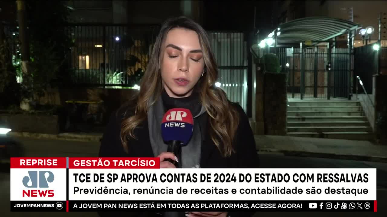 A Jovem Pan News reporter stands outside, speaking into a microphone with the JP News logo. Behind her, a gated building is dimly lit at night in Brazil.
