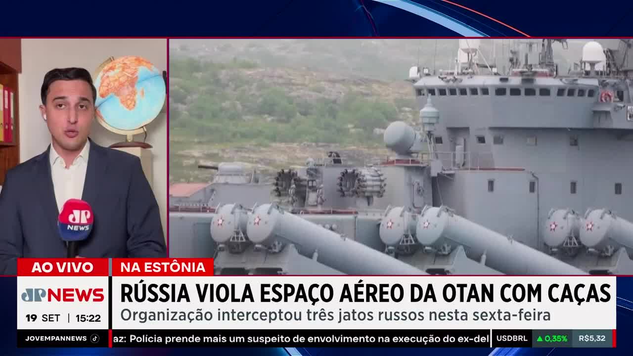 A news anchor from Jovem Pan News in Brazil is reporting live, a globe visible behind him.  Behind the anchor, a gray warship is docked, its missiles prominently displayed.
