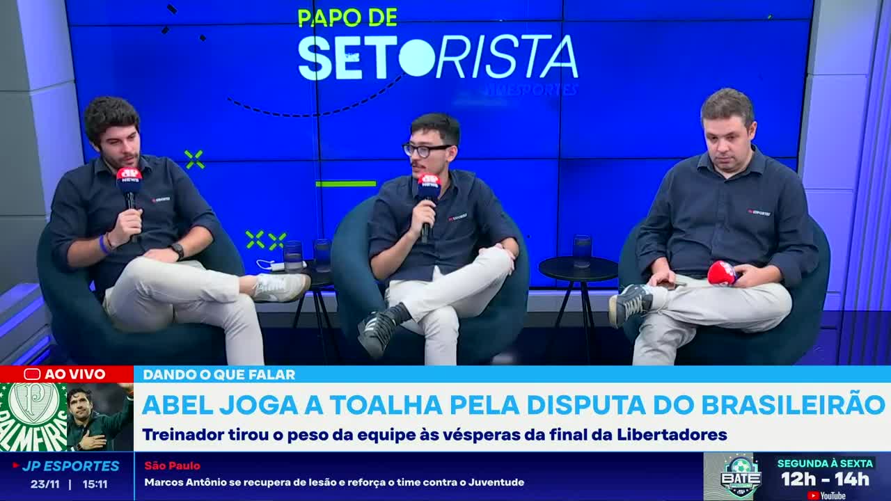 Three men, each holding a microphone, sit in comfortable chairs against a bright blue backdrop. The Jovem Pan News broadcast is discussing Abel's decision in the Brasileirão, with a graphic about the Libertadores final displayed below.
Three men, each holding a microphone, sit in comfortable chairs against a bright blue backdrop. The Jovem Pan News broadcast is discussing Abel's decision in the Brasileirão, with a graphic about the Libertadores final displayed below.