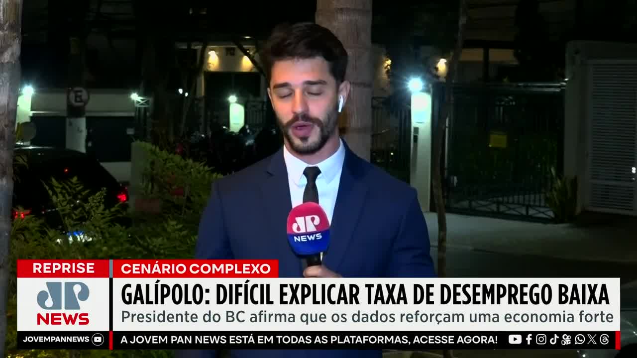 A Jovem Pan News reporter, microphone in hand, speaks into the camera, the backdrop a dark street in Brazil. The chyron reads "Galípolo: Difficult to explain low unemployment rate."
A Jovem Pan News reporter, microphone in hand, speaks into the camera, the backdrop a dark street in Brazil. The chyron reads "Galípolo: Difficult to explain low unemployment rate."