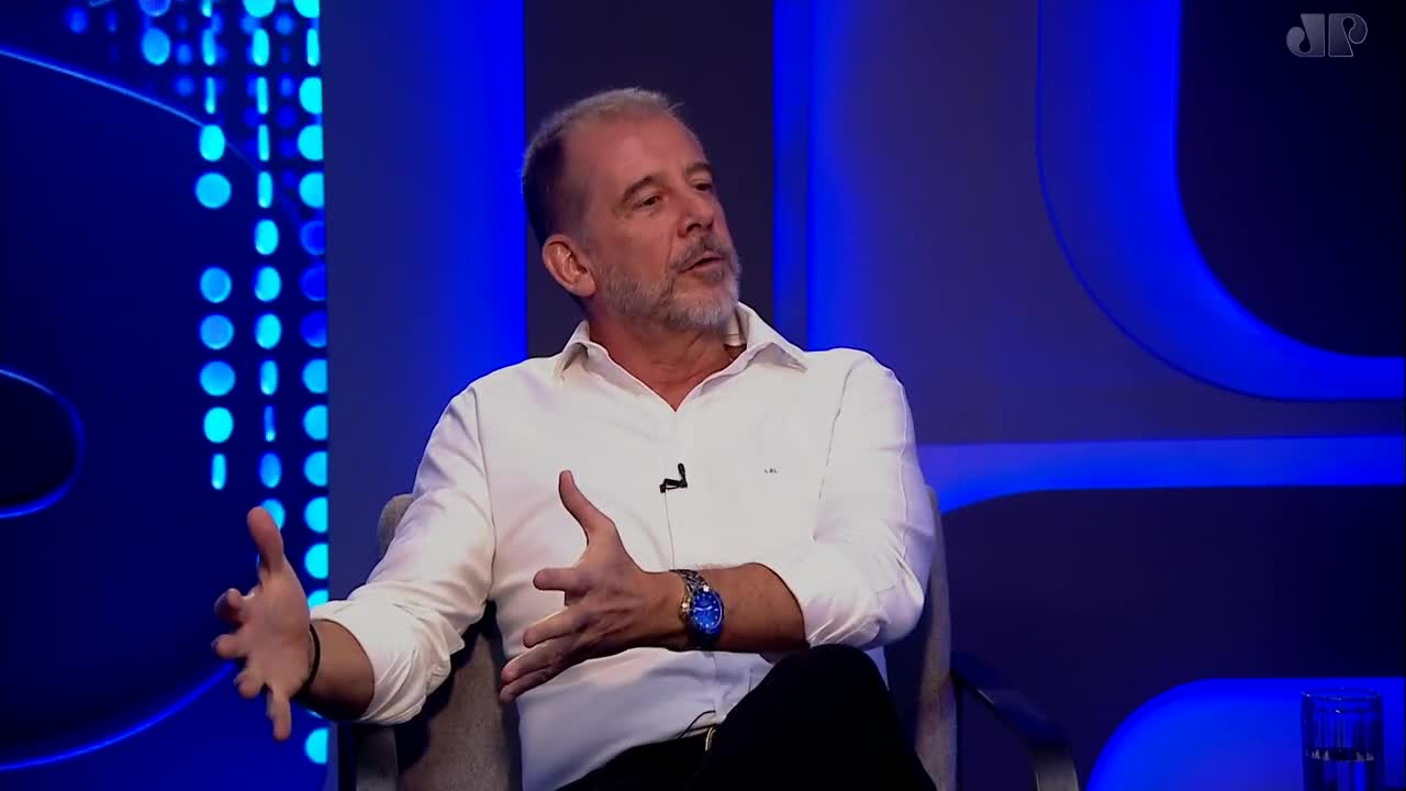 A man in a white shirt gestures with his hands while speaking. The Jovem Pan News logo is visible on the blue backdrop. A man in a white shirt gestures with his hands while speaking. The Jovem Pan News logo is visible on the blue backdrop.