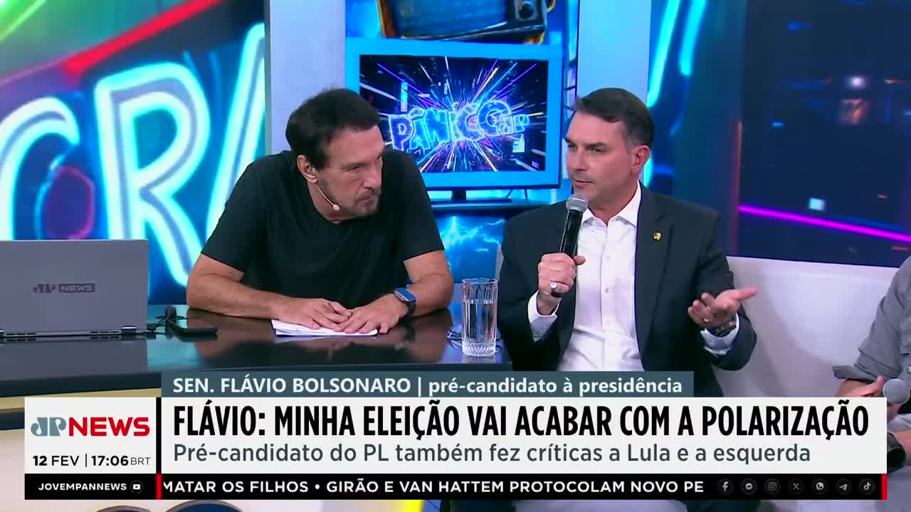 Flávio Bolsonaro, a presidential hopeful, speaks into a microphone while gesturing with his other hand. He sits across from a man in a black t-shirt, both at a table in front of a Jovem Pan News broadcast. Flávio Bolsonaro, a presidential hopeful, speaks into a microphone while gesturing with his other hand. He sits across from a man in a black t-shirt, both at a table in front of a Jovem Pan News broadcast.