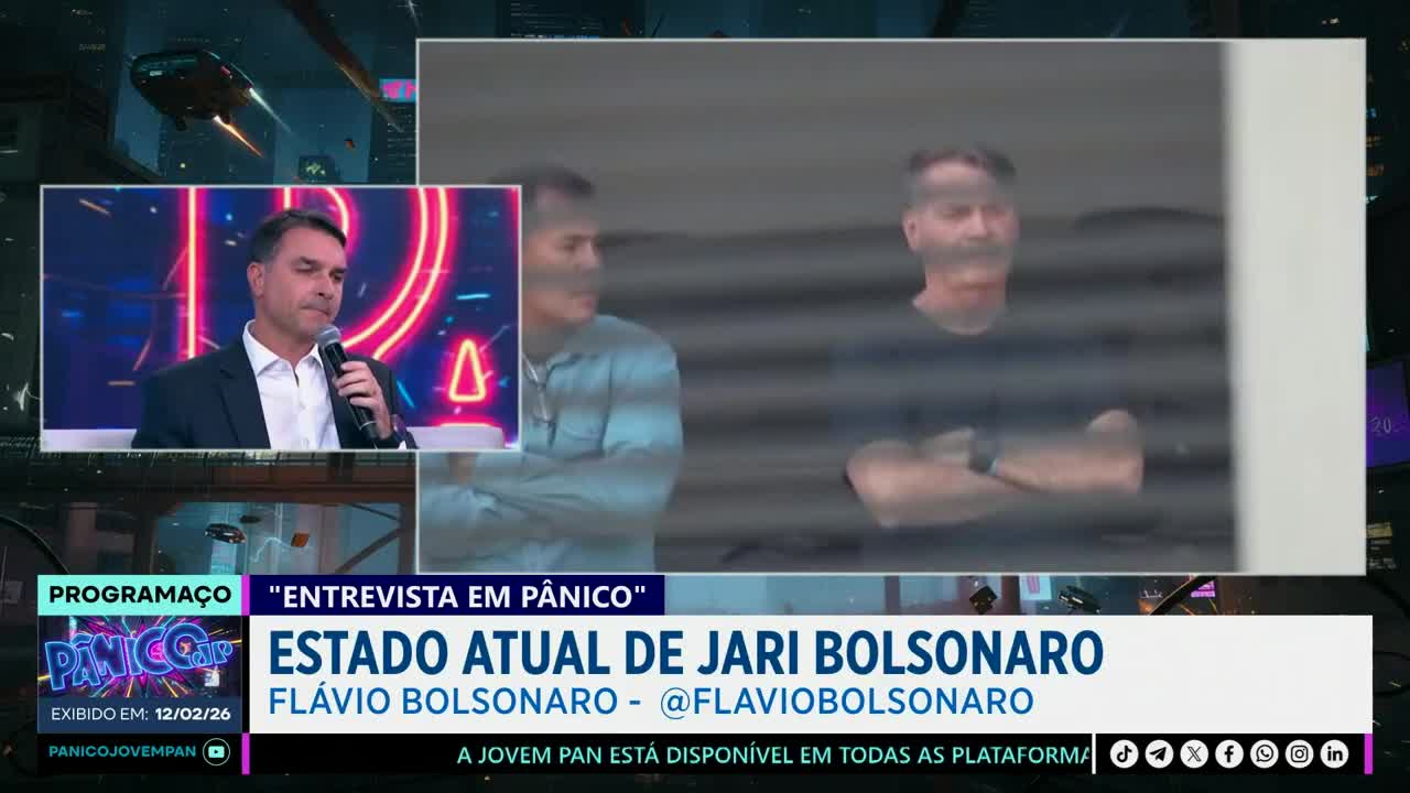 A man in a suit speaks into a microphone on a brightly lit set. Behind him, a man with crossed arms stands in front of a gray, slatted wall. A man in a suit speaks into a microphone on a brightly lit set. Behind him, a man with crossed arms stands in front of a gray, slatted wall.