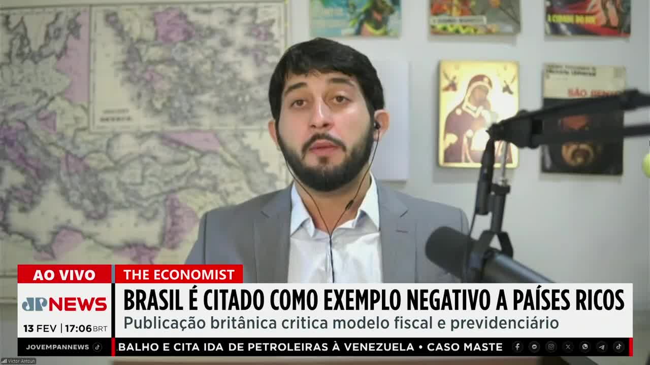 A man with a beard, wearing a suit jacket and a light blue shirt, speaks into a microphone. Behind him, a large map of Europe hangs on the wall, and a news ticker displays "Jovem Pan News" and "Brasil é citado como exemplo negativo a países ricos." A man with a beard, wearing a suit jacket and a light blue shirt, speaks into a microphone. Behind him, a large map of Europe hangs on the wall, and a news ticker displays "Jovem Pan News" and "Brasil é citado como exemplo negativo a países ricos."