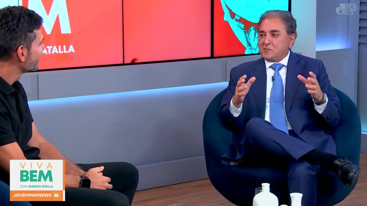 A man in a sharp blue suit gestures with both hands, speaking animatedly on a Jovem Pan News set. Across from him, another man in a black polo shirt listens intently. A man in a sharp blue suit gestures with both hands, speaking animatedly on a Jovem Pan News set. Across from him, another man in a black polo shirt listens intently.