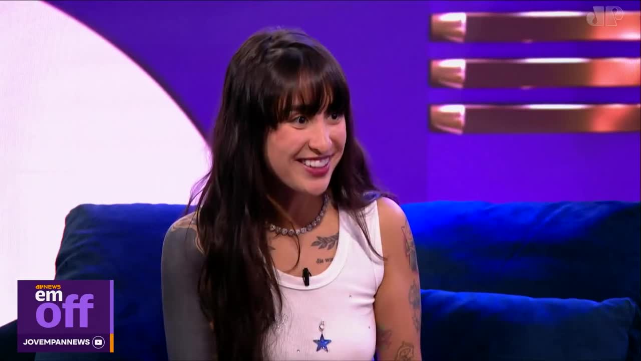 A young woman with dark hair and tattoos smiles broadly while sitting on a blue couch. She's wearing a white tank top and a beaded necklace, appearing on a Jovem Pan News program. A young woman with dark hair and tattoos smiles broadly while sitting on a blue couch. She's wearing a white tank top and a beaded necklace, appearing on a Jovem Pan News program.