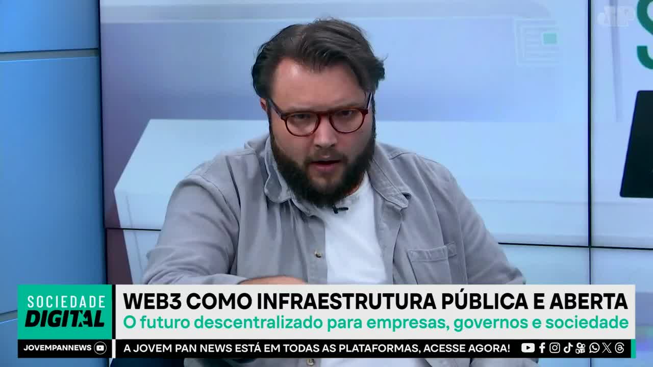 A man with glasses and a beard speaks on a Jovem Pan News program in Brazil. The screen behind him displays text about "WEB3 as public and open infrastructure." A man with glasses and a beard speaks on a Jovem Pan News program in Brazil. The screen behind him displays text about "WEB3 as public and open infrastructure."