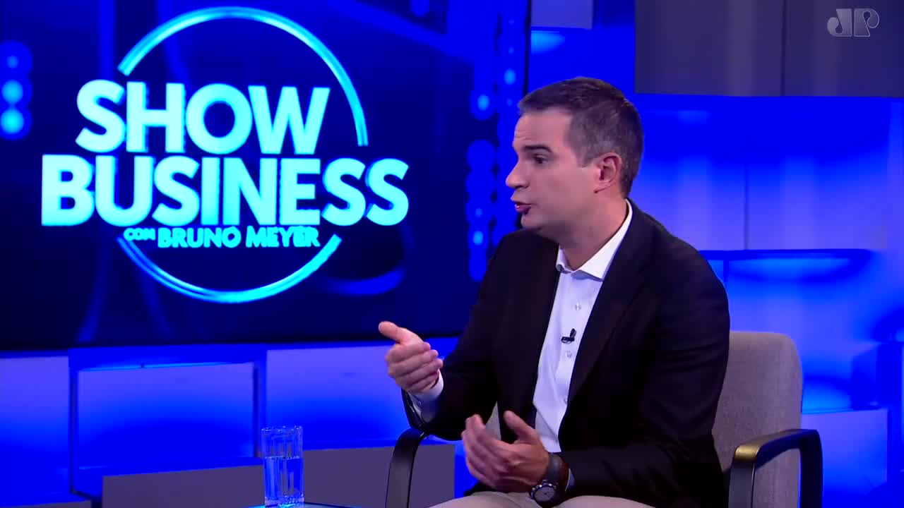 A man in a suit gestures with his hands while speaking. Behind him, the "Show Business" logo glows on a screen, a familiar sight on Jovem Pan News.