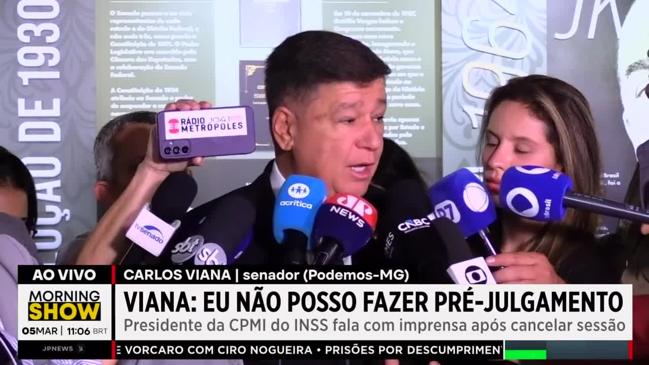 Senator Carlos Viana is speaking into a cluster of microphones, holding up a phone showing the Radio 104.1 Metrópoles logo. A young woman stands beside him, also with microphones pointed at her face.