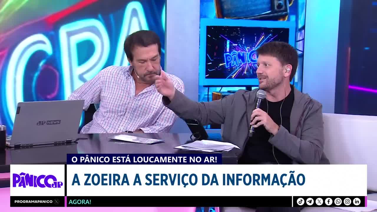 A man in a grey jacket gestures with his right hand while holding a microphone. To his left, another man in a checkered shirt looks down, his hand near his mouth.