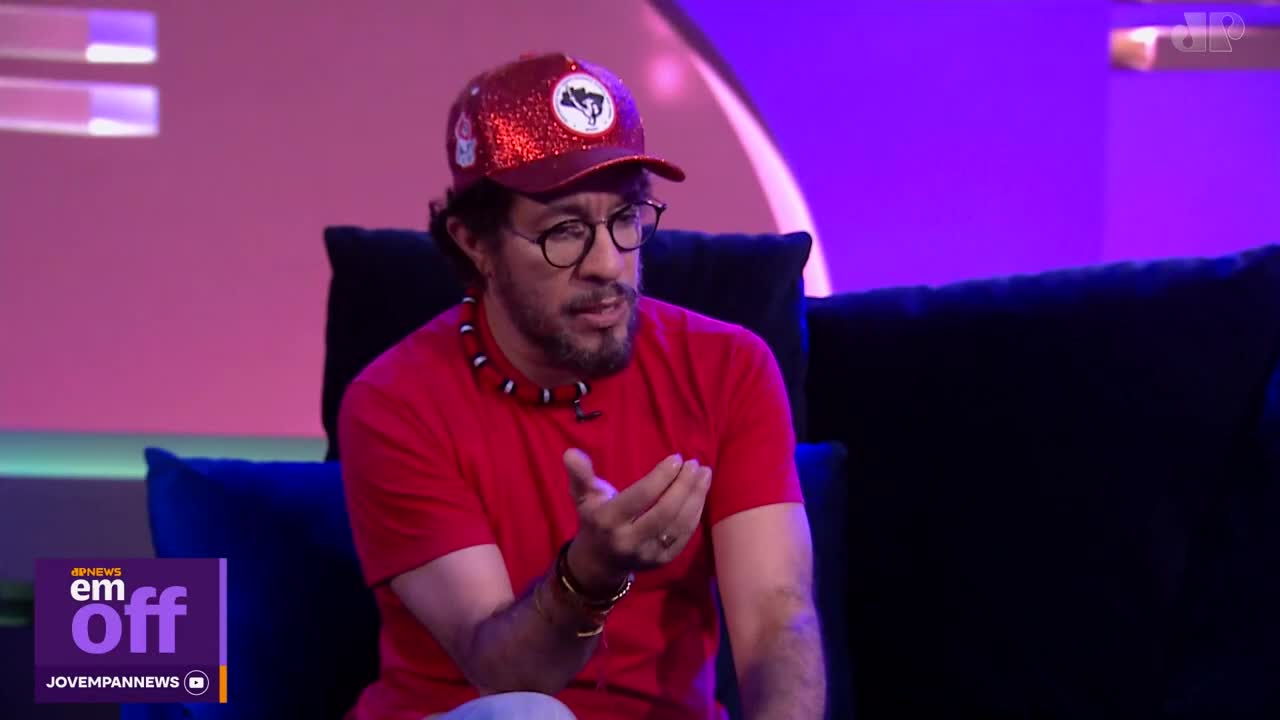 A man in a sparkly red cap and a red shirt gestures with his hand while speaking. He wears a beaded necklace and glasses, sitting on a dark couch in front of a purple backdrop.