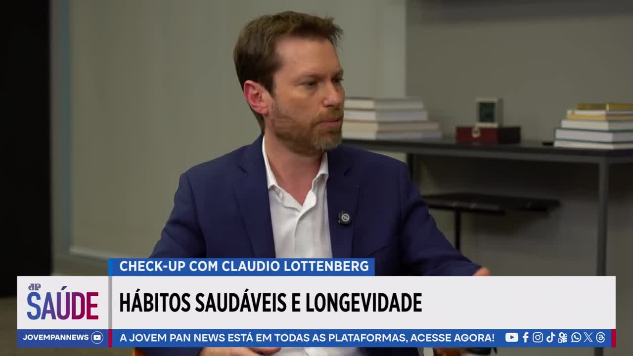 A man in a blue suit jacket and white shirt speaks, his gaze directed slightly off-camera. Behind him, a shelf holds stacks of books and a small decorative box.