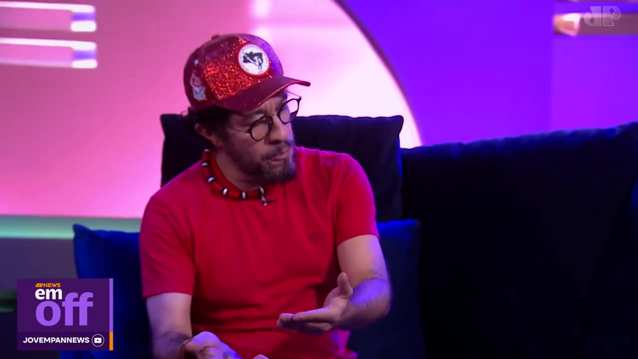 A man in a red shirt and a sparkly red cap gestures with his hands, speaking on a Jovem Pan News program. The studio lights cast a vibrant pink and purple glow around him.