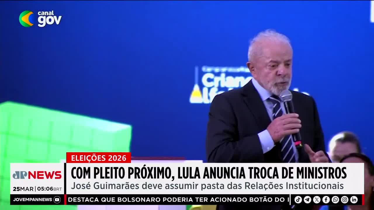 A man with a gray beard, wearing a dark suit and striped tie, speaks into a microphone. Behind him, a blue screen displays text and graphics, including a "Canal Gov" logo and a Jovem Pan News chyron about upcoming elections. A man with a gray beard, wearing a dark suit and striped tie, speaks into a microphone. Behind him, a blue screen displays text and graphics, including a "Canal Gov" logo and a Jovem Pan News chyron about upcoming elections.