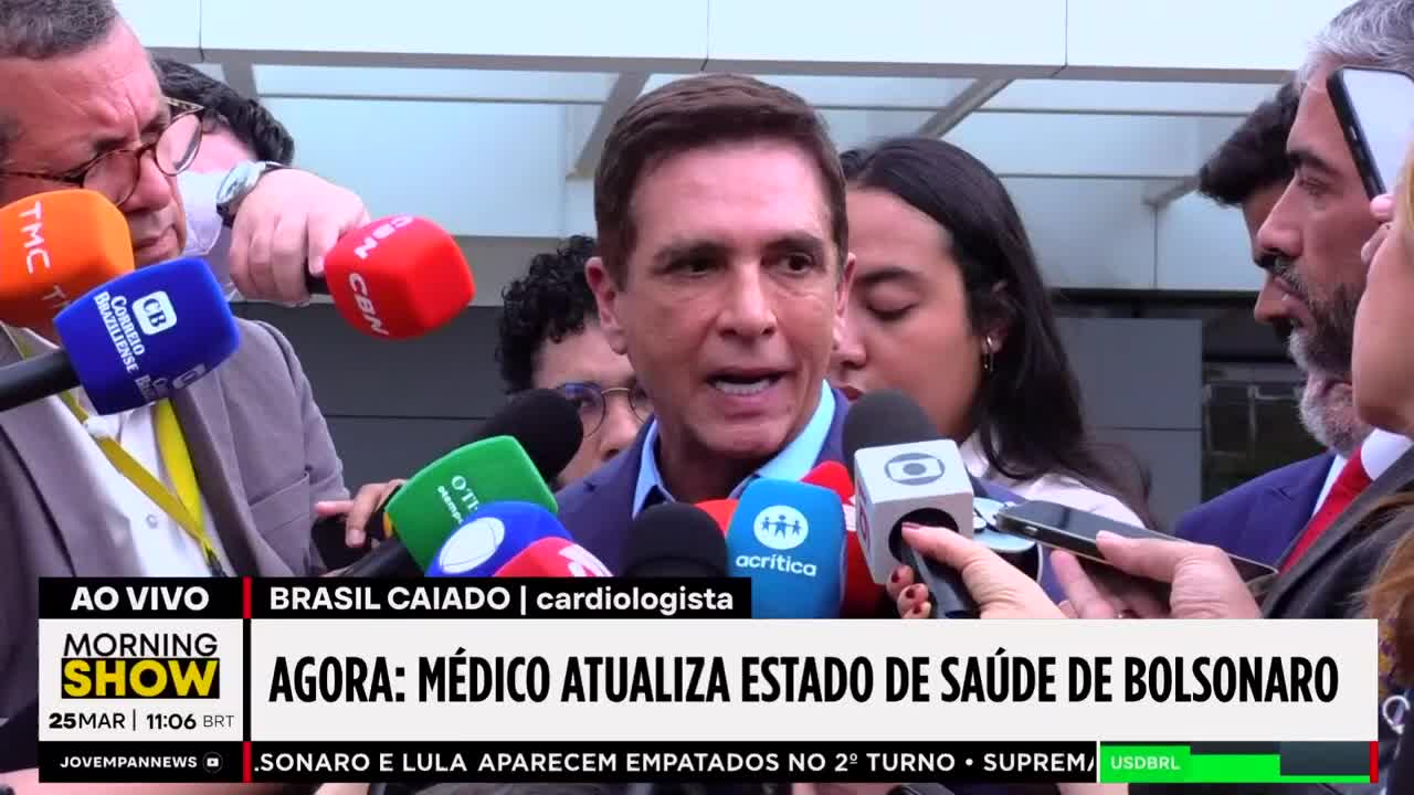A cardiologist speaks into a cluster of microphones, his words likely for Jovem Pan News. Several reporters hold their devices, capturing his statement live from Brazil. A cardiologist speaks into a cluster of microphones, his words likely for Jovem Pan News. Several reporters hold their devices, capturing his statement live from Brazil.