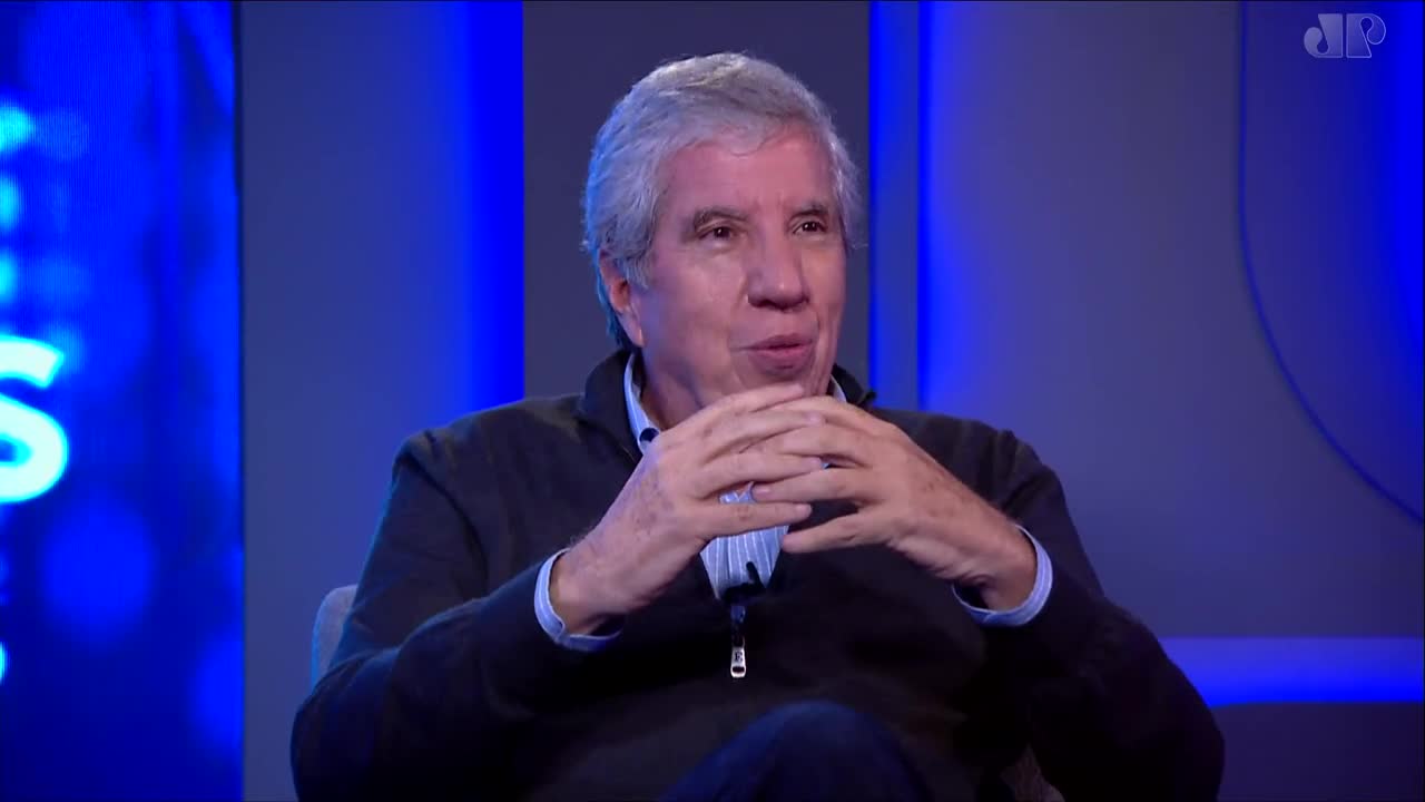 The man's hands are clasped in front of him, his fingers intertwined. He's speaking, his mouth slightly open, against a backdrop of blue lights. The man's hands are clasped in front of him, his fingers intertwined. He's speaking, his mouth slightly open, against a backdrop of blue lights.