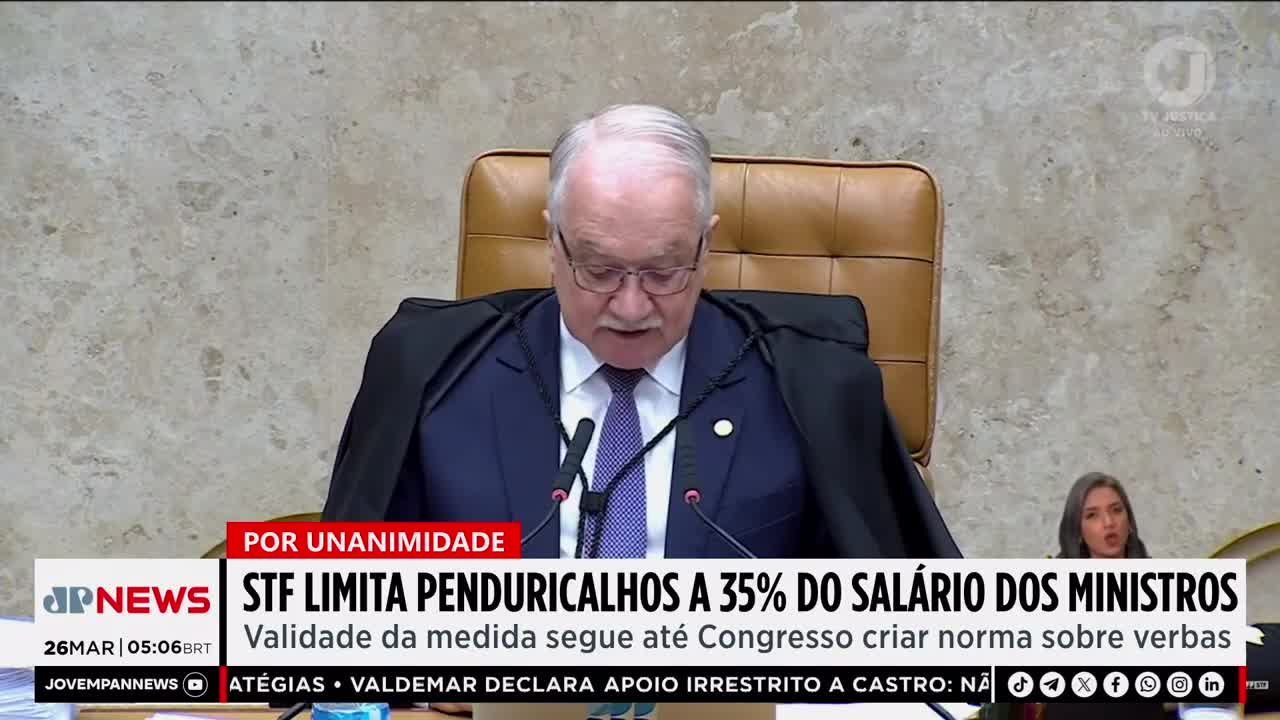 A man in judicial robes speaks into microphones, his face illuminated by the camera's light. A news ticker below displays "Jovem Pan News" and a headline about ministerial salaries. A man in judicial robes speaks into microphones, his face illuminated by the camera's light. A news ticker below displays "Jovem Pan News" and a headline about ministerial salaries.