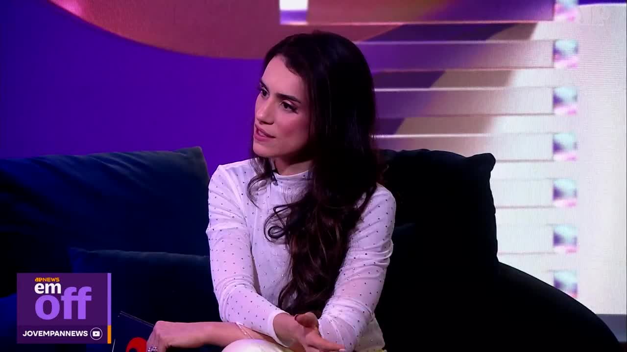 A woman with long dark hair, wearing a white dotted blouse, sits on a dark couch. She's in a studio, likely for a Jovem Pan News program, with a purple backdrop and a screen displaying horizontal bars. A woman with long dark hair, wearing a white dotted blouse, sits on a dark couch. She's in a studio, likely for a Jovem Pan News program, with a purple backdrop and a screen displaying horizontal bars.
