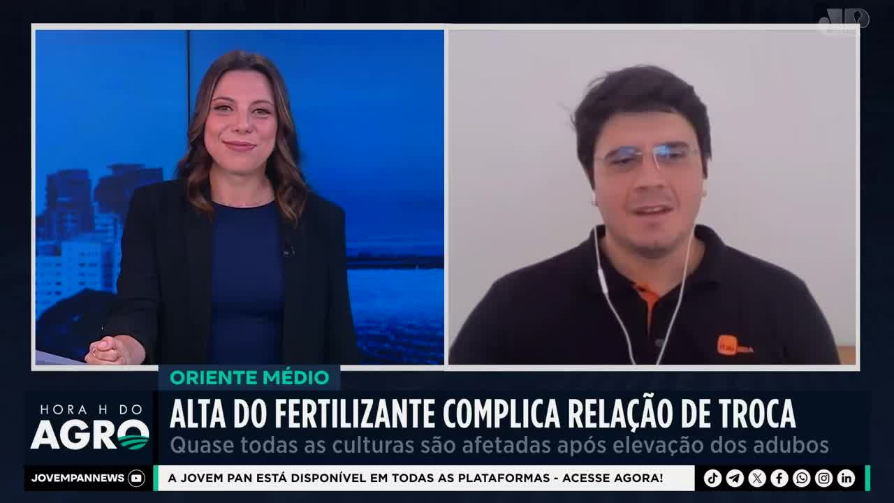 A woman smiles from a studio set, while a man in a black polo shirt appears on a split screen, discussing agricultural news for Jovem Pan. The text at the bottom of the screen indicates a segment on rising fertilizer costs impacting crops in Brazil. A woman smiles from a studio set, while a man in a black polo shirt appears on a split screen, discussing agricultural news for Jovem Pan. The text at the bottom of the screen indicates a segment on rising fertilizer costs impacting crops in Brazil.