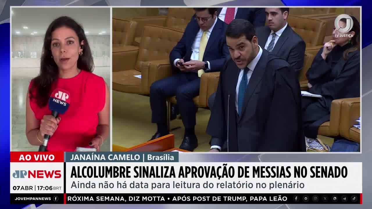 A man in a dark robe speaks with conviction, gesturing with his hand. Behind him, other figures in formal attire sit in tiered seating. To the left, a reporter in a vibrant red dress holds a microphone, addressing the camera.