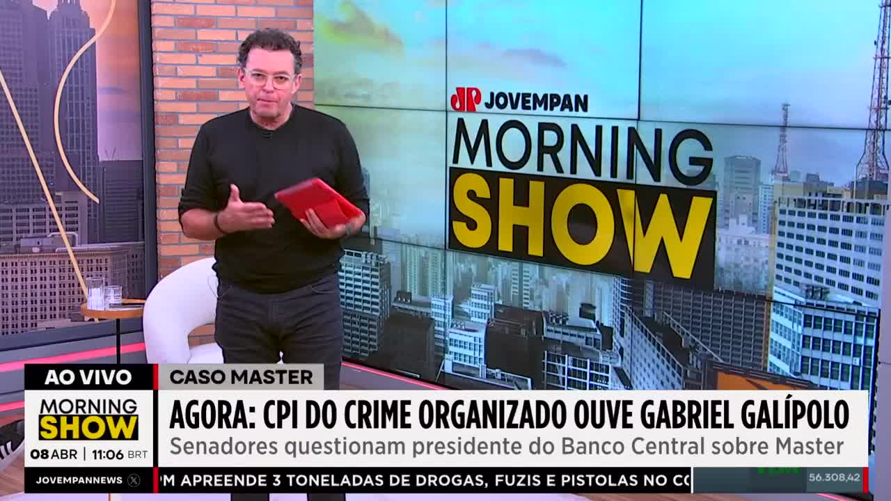 A man in a black shirt stands holding a red tablet, gesturing with his left hand. Behind him, the Jovem Pan Morning Show logo is displayed over a city skyline.
