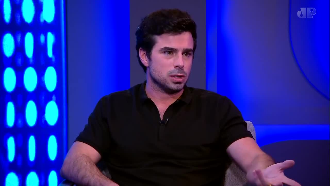 A man in a black polo shirt gestures with his right hand while speaking. Behind him, a wall glows with blue lights.