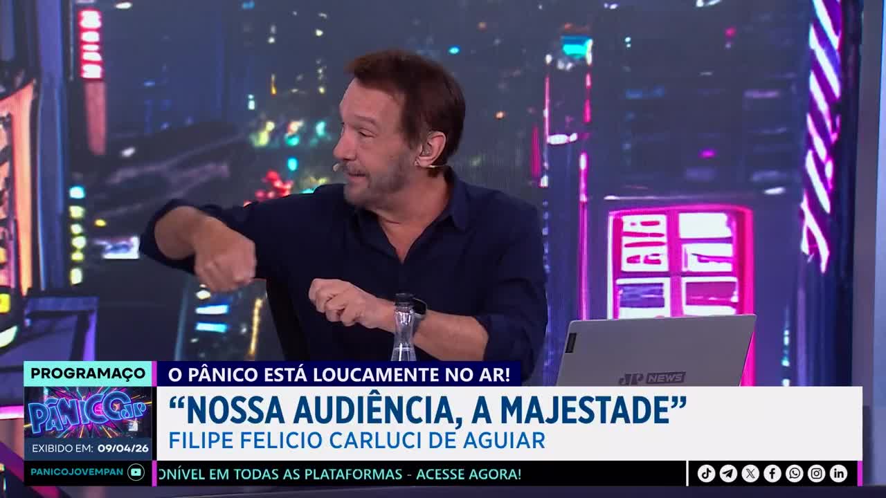 A man in a dark shirt gestures with his right hand, his left fist clenched. Behind him, a screen displays the Jovem Pan News logo and text in Portuguese.