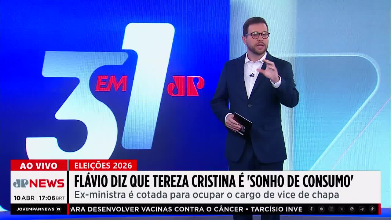 A man in a dark suit stands before a large blue screen displaying "31 EM JP" and election graphics. He gestures with his hands as he speaks, holding a tablet in his left arm.