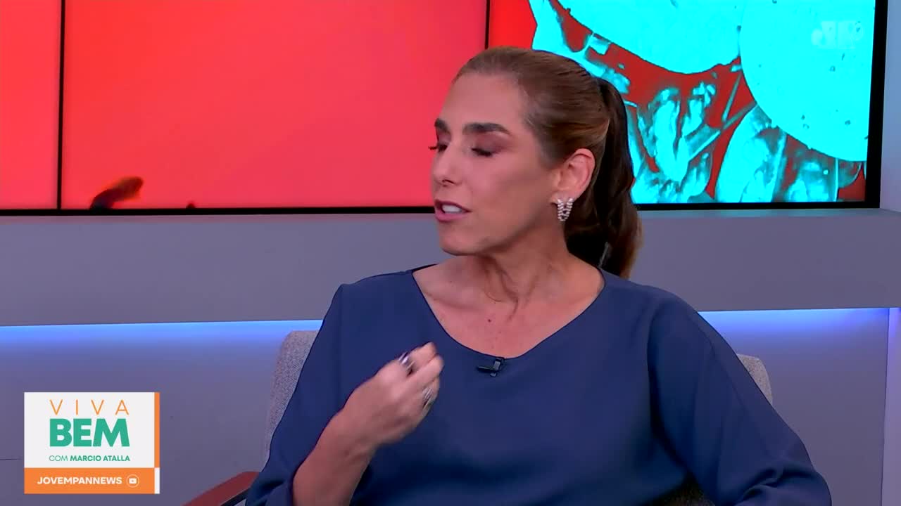 A woman in a blue top speaks on a talk show set, her hand gesturing. Behind her, a screen displays abstract red and blue graphics, with a small, dark shape moving across the red section.