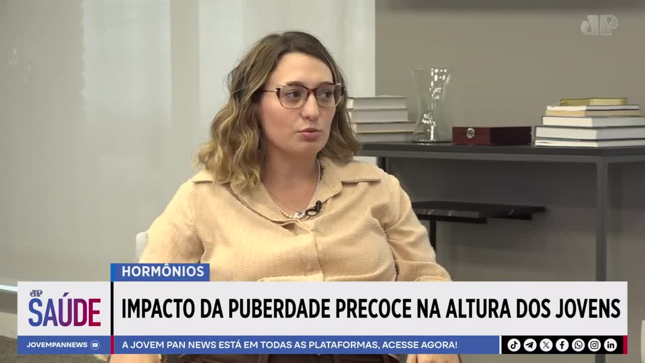 A woman with blonde, wavy hair and glasses speaks into a microphone, her light brown shirt buttoned at the collar. Behind her, a shelf holds stacks of books and a decorative vase. A graphic overlay reads "SAÚDE" and "IMPACTO DA PUBERDADE PRECOCE NA ALTURA DOS JOVENS" from Jovem Pan News.