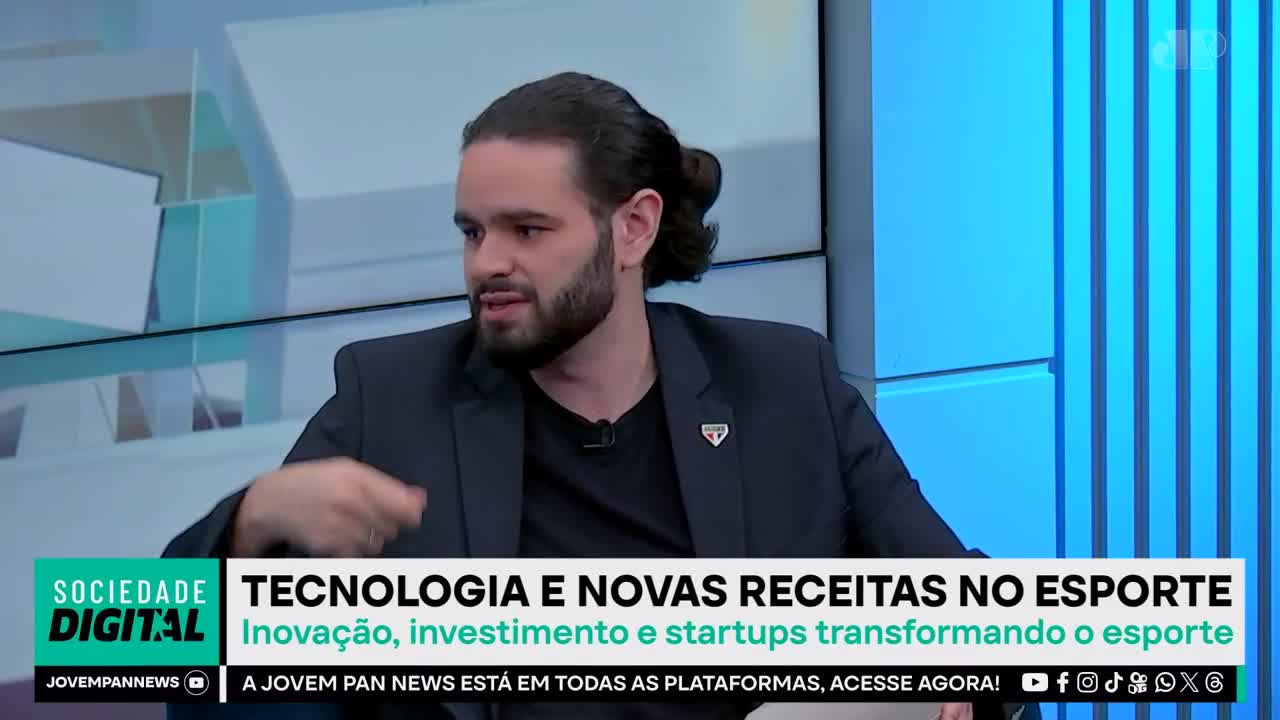 A man with his hair pulled back in a bun gestures with his left hand while speaking on a Jovem Pan News broadcast. The screen displays "Tecnologia e Novas Receitas no Esporte," discussing innovation and startups in sports.