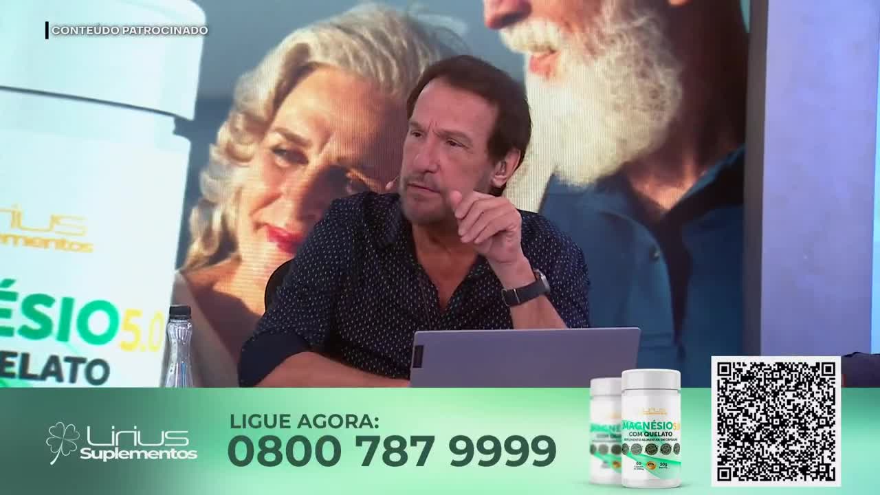 A man with a serious expression leans forward, his hand touching his chin. Behind him, a large screen displays a woman with gray hair and a man with a white beard, likely from a Jovem Pan News segment.