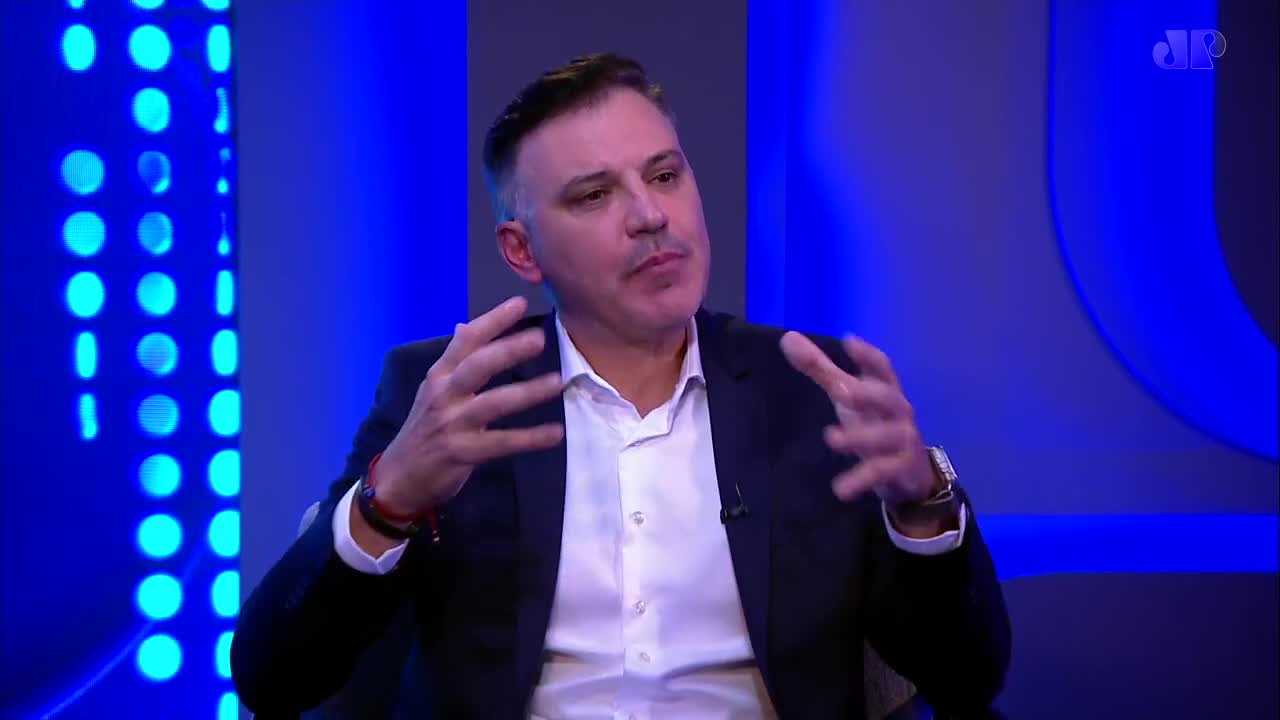 A man in a dark suit and white shirt gestures with both hands, speaking animatedly on a Jovem Pan News broadcast. The background is a vibrant blue, with a pattern of illuminated circles on the left.