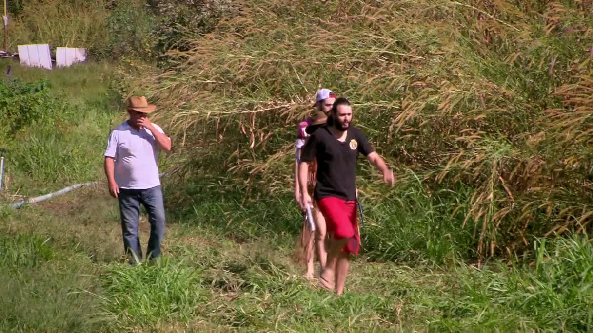 A man in a black shirt and red shorts strides through tall grass, followed by two others. Nearby, a man in a cowboy hat looks on, hand at his face.
