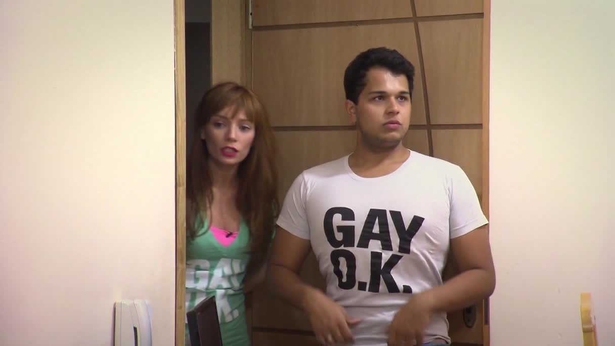 A woman with long red hair and a man in a "GAY O.K." t-shirt stand in a doorway, looking off to the side. They seem to be in a scene for Brazilian MTV.
