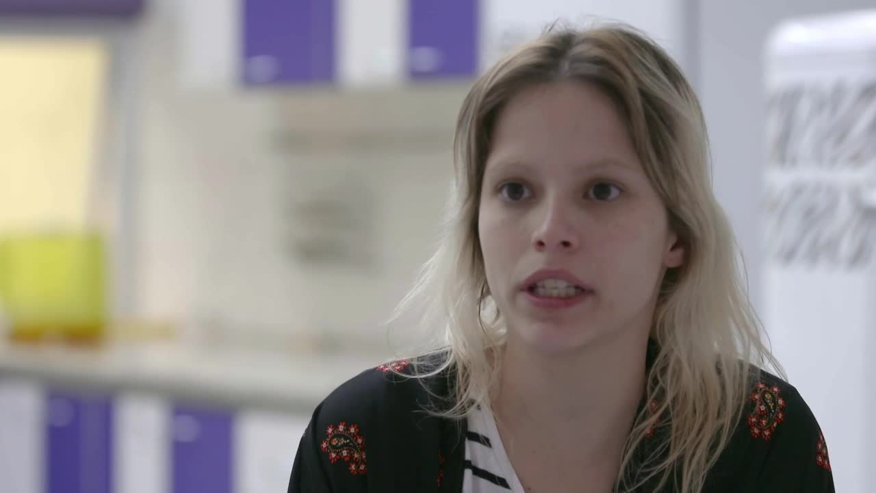 A woman with blonde hair is speaking, her mouth moving as she forms words. She's wearing a black patterned shirt with a white and black striped undershirt.
