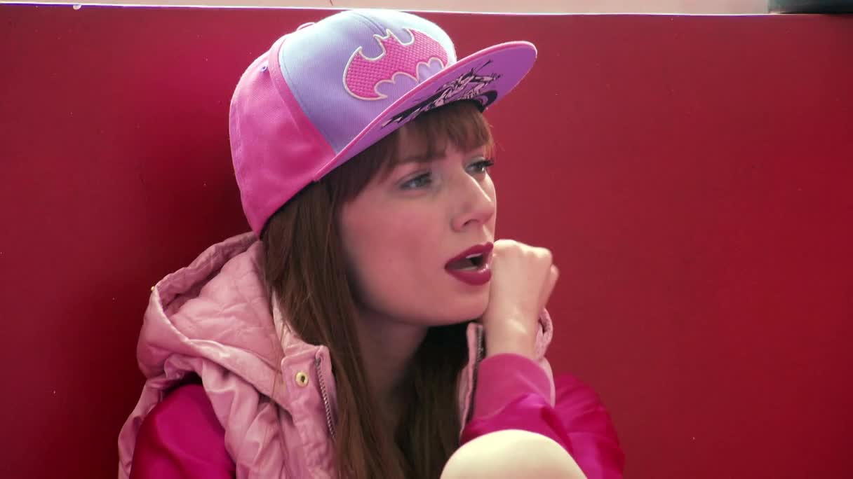 She's leaning back, wearing a pink Batman cap and jacket, her lips slightly parted as if she's about to speak. The red backdrop behind her makes the whole scene pop, like something from an MTV Brazil set.

