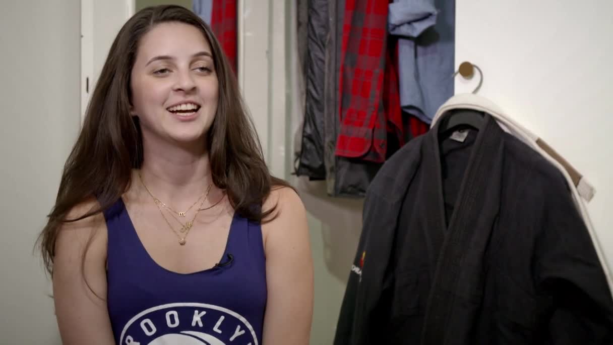 A woman in a Brooklyn tank top is talking, her smile wide. Behind her, a black Brazilian Jiu-Jitsu gi hangs on a hook.
