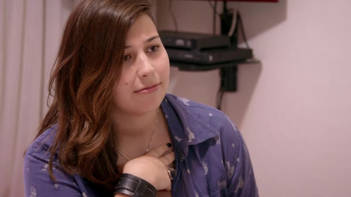The woman in the blue shirt is looking off to the side, her hand resting on her chest. She's wearing a thick black wristband and a silver necklace.
